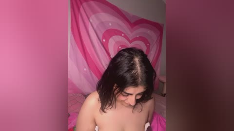 Media: Video of a young woman with shoulder-length dark hair, fair skin, and small breasts, topless, in a room with pink heart-patterned wall hanging, sitting on a pink bedspread.