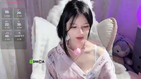 Media: Video of a young Asian woman with black hair, wearing a pastel floral kimono, sitting on a plush white chair with a teddy bear. She has a pink pacifier in her mouth.