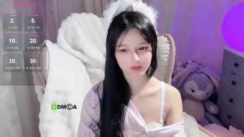 Media: A video of a young East Asian woman with long black hair, wearing a white feathered headpiece, a light pink satin robe, and white lingerie. She sits on a plush, white bed in a softly lit room, with a plush toy beside her.