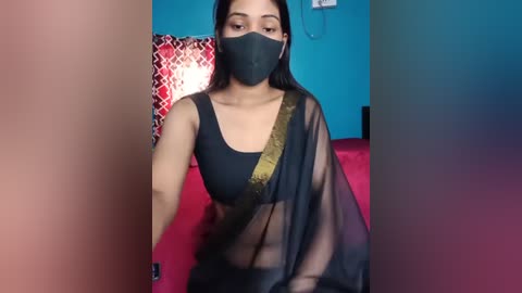 Media: Video of a South Asian woman with medium skin tone, long black hair, wearing a black mask, black bra, and see-through black sari with gold trim, sitting on a red couch against a teal wall.