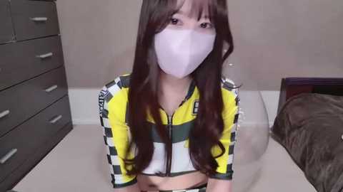 Media: Video of an Asian woman with long, dark hair, wearing a yellow and black sports jacket, white face mask, and sitting on a beige floor in a simple bedroom.