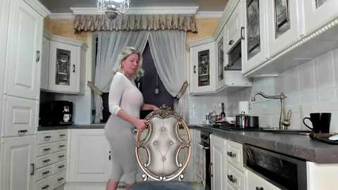 Media: Video of a woman with blonde hair, wearing a white blouse and gray skirt, pushing a decorative chair in a modern kitchen with white cabinets, stainless steel appliances, and a window with sheer drapes.