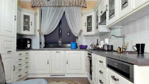 Media: Video of a modern kitchen with white cabinets, stainless steel appliances, and a black countertop. A blue bowl sits on the counter next to a microwave and a sink with a window adorned with sheer curtains.