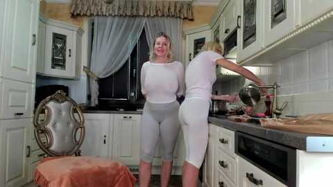 Media: A video shows two women in matching white, see-through tops and leggings, one washing dishes, the other standing in a cozy kitchen with white cabinets, a floral window curtain, and a rustic chair.