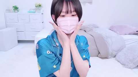 Media: A video of an Asian woman with short black hair, wearing a blue shirt and pink mask, covering her face with both hands, in a bright, minimalistic bedroom with white furniture and fluffy pillows.