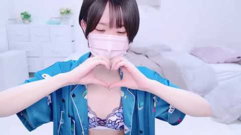 Media: A video of an Asian woman with short black hair, wearing a blue shirt, pink face mask, and heart hands, in a white bedroom with a bed and dresser.