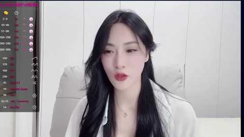 Media: Video of a young Asian woman with long black hair, fair skin, and red lipstick, sitting indoors against a white wall, wearing a white shirt.