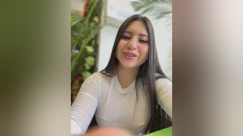 Media: Video of a young Latina woman with long dark hair, wearing a sheer white long-sleeve top, smiling at the camera in a lush indoor setting with greenery and a white wall.