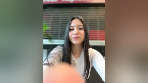 Media: Video of a young Asian woman with long black hair and light skin, wearing a white long-sleeve top, seated indoors with blurred background, possibly in a fast-food restaurant.