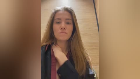 Media: Video of a young Caucasian woman with long, straight brown hair, wearing a maroon top and dark jacket, standing in a beige-tiled hallway.