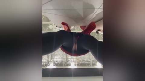 Media: Video of a muscular male superhero in a red suit, white mask, and black boots, leaping towards the camera, legs spread, revealing his bulging crotch, in a modern indoor setting with a glass ceiling and geometric light fixtures.