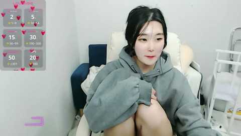 Media: Video of a young East Asian woman with long black hair, wearing a grey hoodie, sitting on a white chair in a clinical setting. Background includes medical equipment and a heart-themed countdown clock.