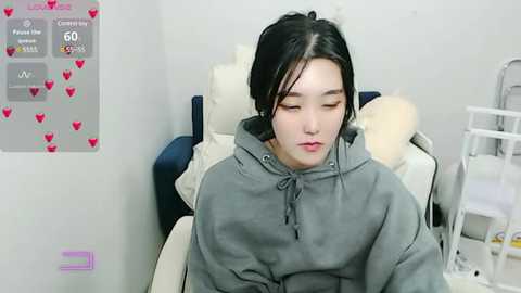 Media: Video of a young Asian woman with long black hair, wearing a gray hoodie, sitting on a white couch in a plain, light-colored room. A medical chart with heart icons and statistics is visible on the left wall.