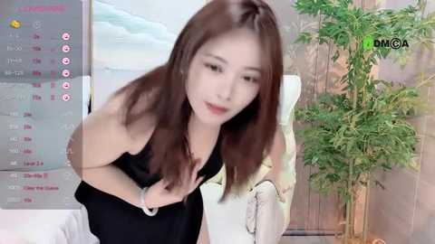 Media: Video of an Asian woman with straight brown hair, wearing a black dress, adjusting it, in a modern indoor setting with a green plant and a calendar displaying dates.