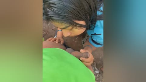 Media: A young girl with long black hair and a turquoise shirt is holding a small, erect penis, likely a child's, outdoors on a dirt ground.