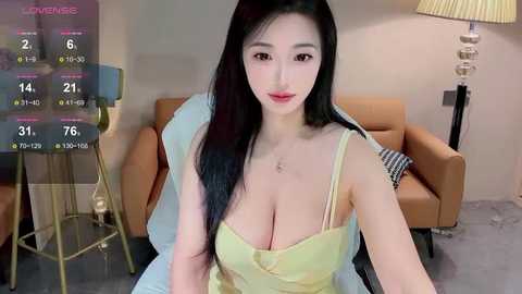 Media: A video of a young East Asian woman with long black hair, fair skin, and a slender figure, wearing a yellow spaghetti strap top, sitting on a beige couch in a modern living room.