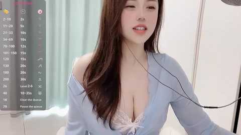 Media: Video of an East Asian woman with long, straight brown hair, fair skin, wearing a light blue off-shoulder top that reveals a lacy white bra, sitting in a sterile hospital room with a monitor in the background.