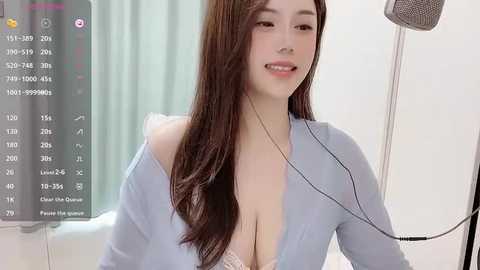 Media: Video of a smiling, fair-skinned Asian woman with long, straight brown hair, wearing a light blue cardigan and a lacy bra, sitting in a modern room with a gray screen displaying a video call.