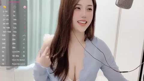 Media: Video of an Asian woman with long brown hair, wearing a low-cut blue top, smiling, sitting in a white room with a microphone.