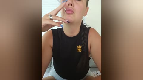 Media: Video of a young woman with medium skin tone and dark hair, wearing a black sleeveless top with a gold crown logo, making a pouty face, fingers on her cheek.