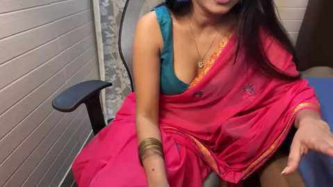 Media: Video of a South Asian woman with medium skin tone, wearing a red sari with a teal blouse, sitting on a black office chair. She has long black hair and is in a small, tiled room with white walls.