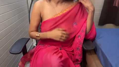 Media: Video of a woman with medium skin tone, wearing a bright pink sari, sitting in a chair, holding her breast. Background shows a tiled wall and blue bedsheet.