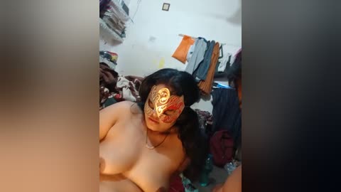 Media: Video of a topless woman with medium breasts, wearing a mask with a tribal design, in a cluttered bedroom with clothes hanging on a rack, a bed, and a desk.