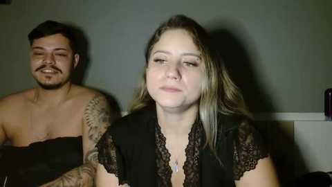 Media: Video of a woman with light skin and long, blonde hair, wearing a black lace top, sitting beside a man with short dark hair and a beard. Both are shirtless, the man with tattoos on his arm. Background is dimly lit, gray wall.