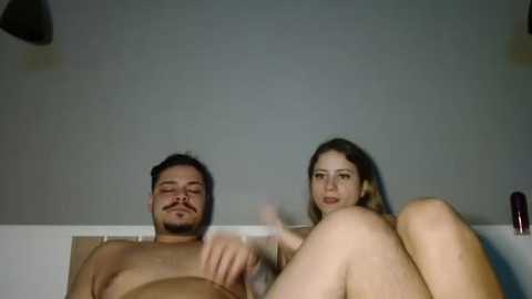 Media: Video of a nude, overweight man and a nude woman, both with light skin and dark hair, lying on a bed in a dimly lit room with grey walls.