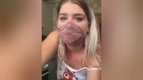 Media: A video of a young woman with light skin and blonde hair, wearing a red sequined face mask and a white tank top with a cartoon bear design. She is indoors, possibly in a kitchen.