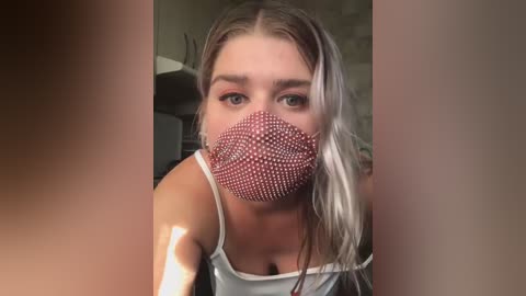 Media: Video of a young woman with light skin, long blonde hair, wearing a red polka-dot face mask and a white tank top. Background shows blurred kitchen appliances.