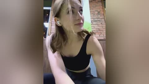 Media: Video of a young woman with light skin and straight, shoulder-length blonde hair, wearing a black sports bra and black leggings, sitting in a modern, dimly lit room with exposed brick walls.