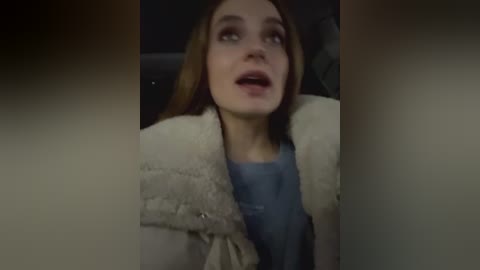 Media: Video of a young woman with long brown hair, wearing a light-colored fur coat over a blue shirt, looking surprised with open mouth, indoors at night.