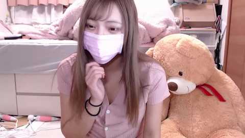 Media: A video of an Asian woman with long brown hair, wearing a white face mask and a light pink button-up shirt, kneeling beside a large stuffed teddy bear in a cluttered bedroom.