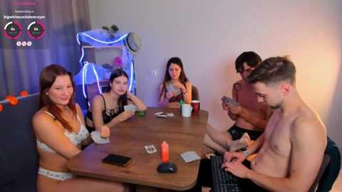 Media: Video of six young adults, three women and three men, in casual attire, sitting around a wooden table in a dimly lit room, playing a card game.