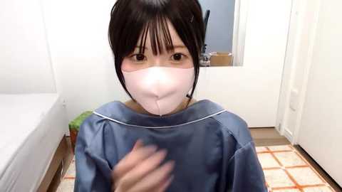 Media: Video of an Asian woman with short black hair, wearing a light blue gown and a pink face mask, standing indoors near a white door.