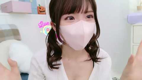 Media: Video of a young Asian woman with long brown hair, wearing a white face mask and pink earrings, in a white robe, standing in a pastel-colored room with a pumpkin decoration on the wall.