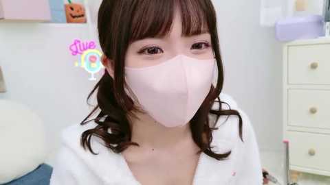 Media: Video of an East Asian woman with long brown hair, wearing a pink surgical mask and white robe, standing in a pastel-colored, child-friendly room with a pumpkin decoration.