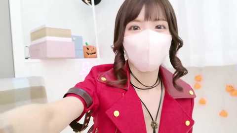 Media: A video of an Asian woman with long brown hair and a white mask, wearing a red jacket with gold buttons, taking a selfie in a room with Halloween decorations.