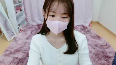 Media: Video of an Asian girl with long black hair, wearing a white knit cardigan and pink face mask, sitting on a purple shaggy rug in a room with white walls and light wooden floor.