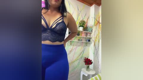 Media: Video of a curvy woman with medium skin tone, wearing a dark blue lace bra and high-waisted blue pants. She stands in a brightly lit room with colorful wall art, a potted plant, and a vase of red flowers on a white shelf.