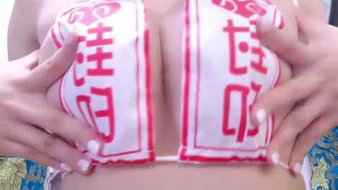 Media: Video of a woman's large, round breasts, partially covered by her hands, with a white bikini top featuring red kanji characters. Background includes a blue and green patterned fabric.