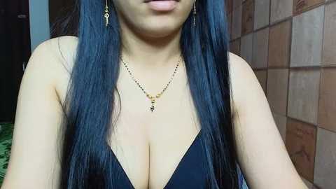 Media: Video of a woman with long, straight black hair, fair skin, and a deep neckline revealing ample cleavage. She wears gold earrings and a gold necklace. Background includes tiled walls and a partially visible door.