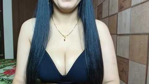 Media: Video of a woman with long, straight, dark blue hair, wearing a low-cut, dark blue, sleeveless top that accentuates her ample cleavage. She has a light skin tone and wears a delicate gold necklace. The background features a tiled wall and a green-patterned rug.