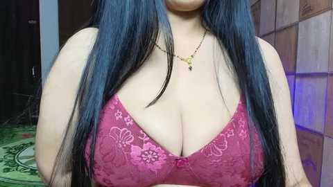 Media: A video of a woman with long, blue-black hair and fair skin, wearing a pink lace bra that accentuates her large breasts, set against a background of a wooden door and a patterned rug.