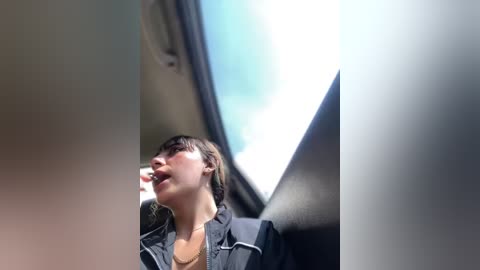 Media: A video of a young woman with short, dark hair, wearing a black jacket and a necklace, standing inside a car with a large, sunlit circular window in the background.