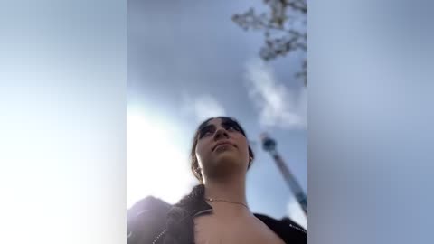 Media: A video capturing a young woman from a low angle, looking upwards with a serene expression. She has medium skin tone and dark hair, wearing a black jacket, against a bright sky with clouds.