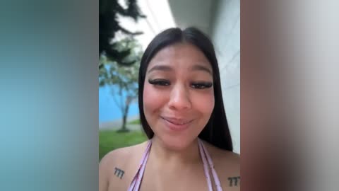 Media: Video of a young woman with long black hair, light brown skin, and a slight smile. She wears a pink halter top and has tattoos on her shoulders. Background features a blue sky, green grass, and a building.