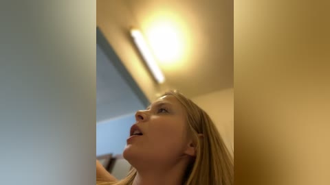 Media: A video of a blonde woman with fair skin, wearing a brown top, gazing upward at a bright ceiling light. The background is blurred, with beige walls.