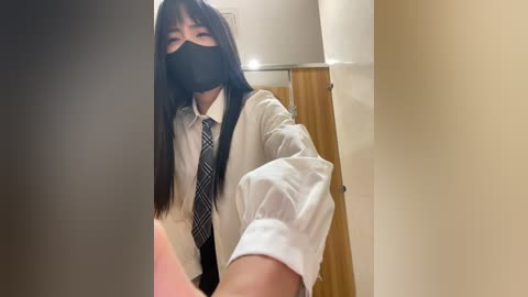 Media: A video shows an East Asian woman with long black hair, wearing a white shirt, black tie, black face mask, and a black mask. She is taking a selfie in a bathroom with beige walls and a wooden door.